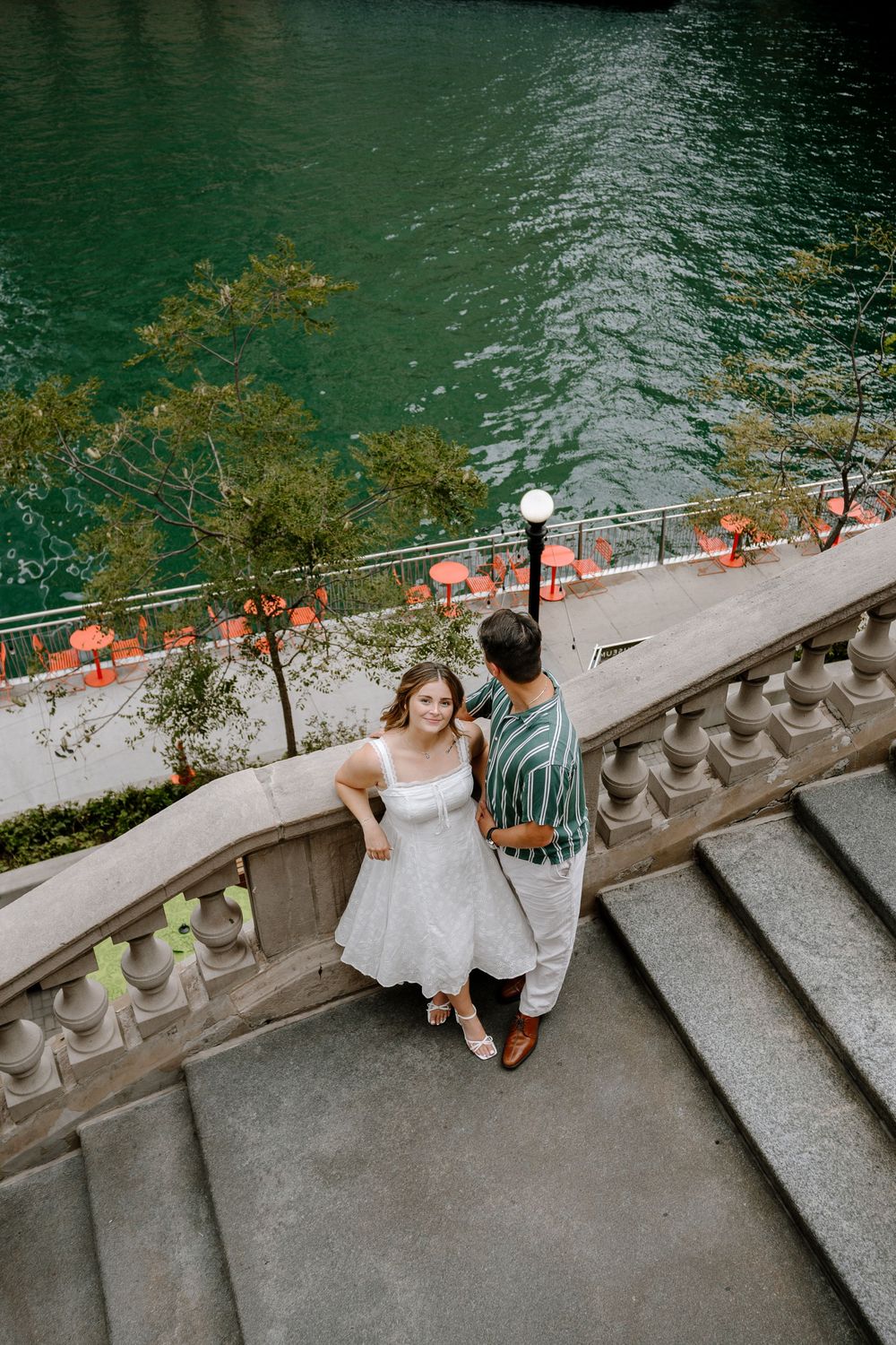 Couple poses on stone steps by Chicago River, woman in white dress, man in striped shirt, aerial view.