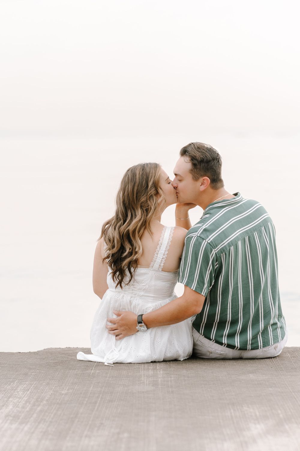 Couple sharing a romantic kiss while sitting on a dock by the water, woman in white dress, man in striped shirt.