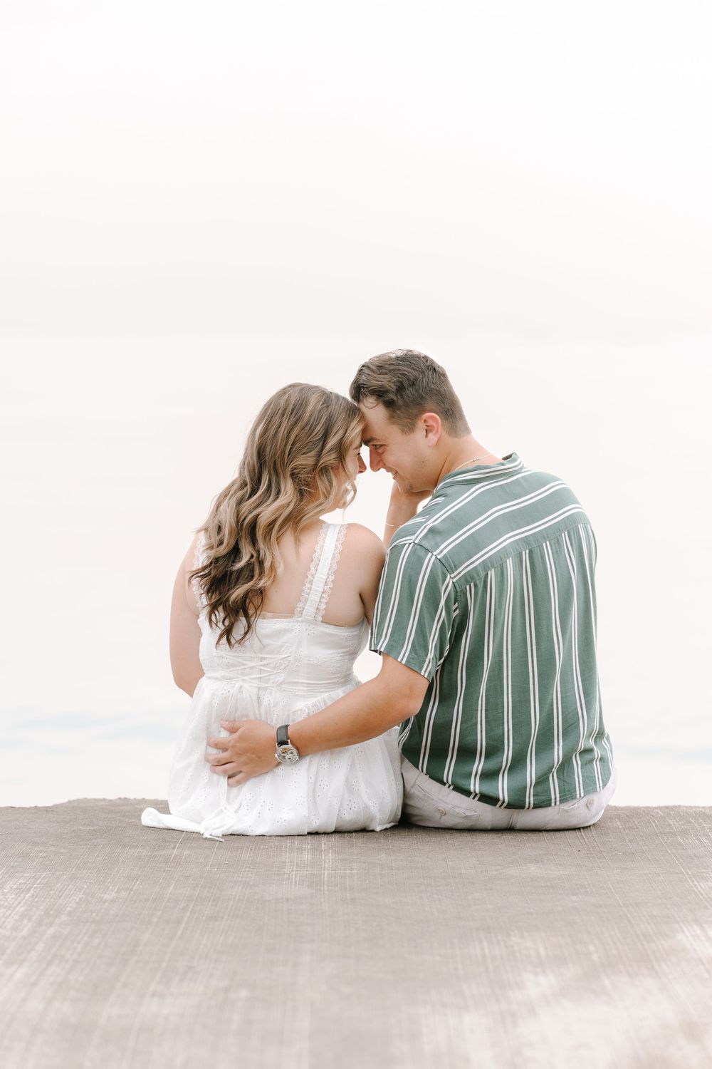 A couple sits together on a dock, touching foreheads romantically with a serene water backdrop.