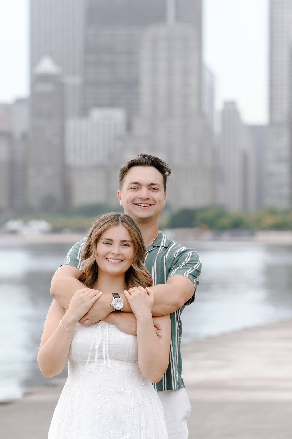 A smiling couple poses on a waterfront with a city skyline backdrop, the man hugging the woman from behind.