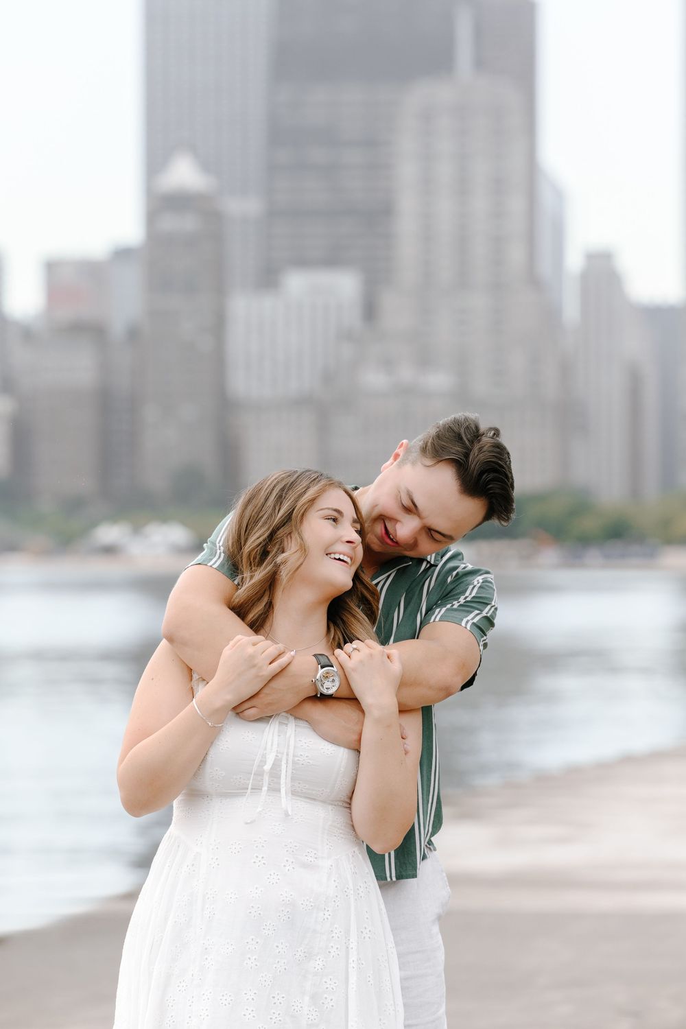 Couple embracing and laughing by a waterfront with city skyline, woman in white dress, man in green striped shirt.