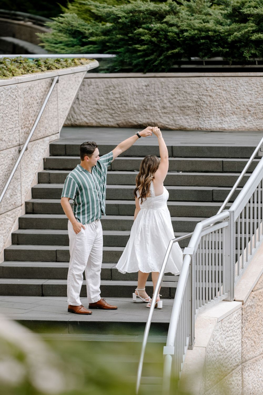 Couple dancing on urban steps, man in green striped shirt, woman spinning in white dress, outdoor setting.