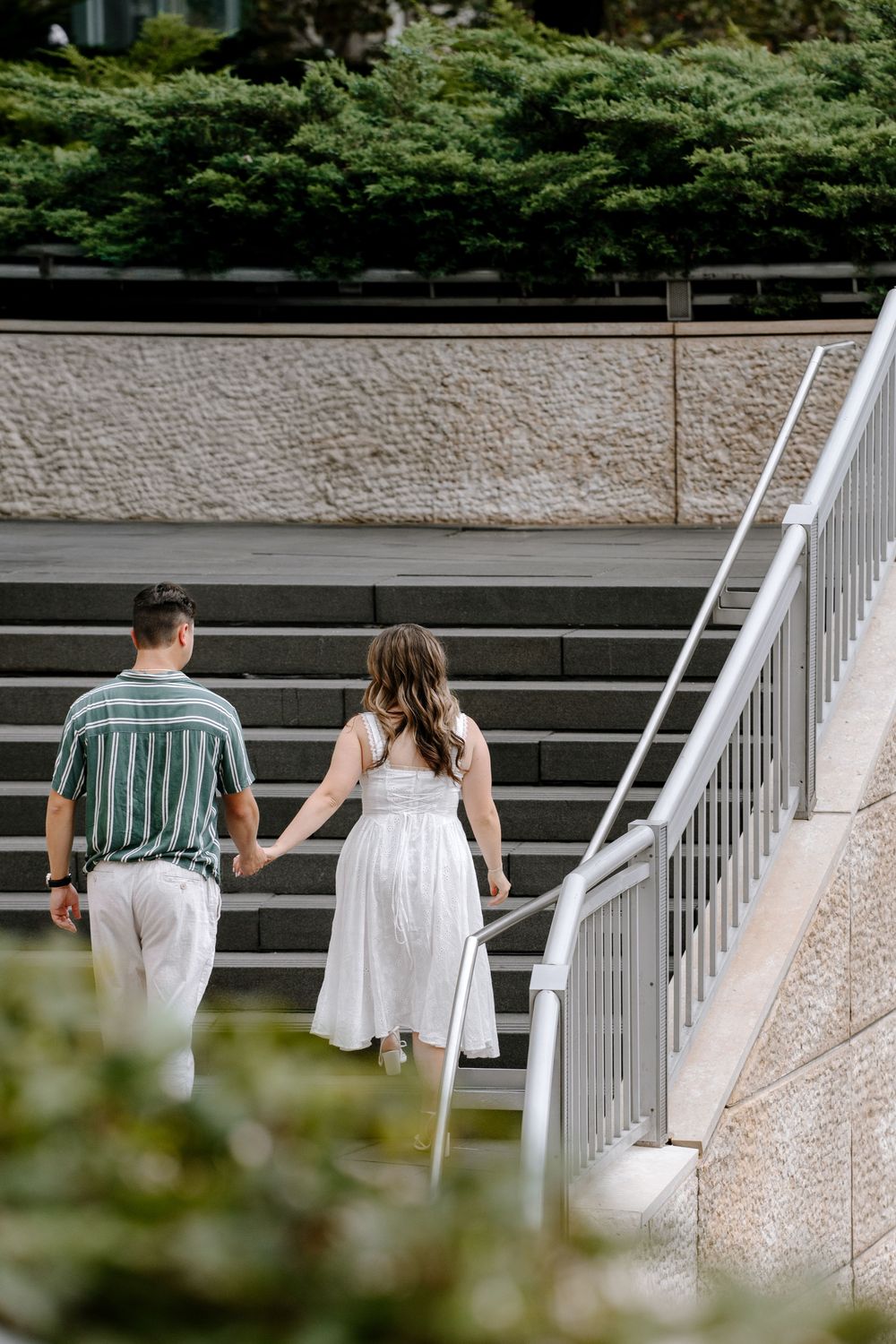 Couple holding hands walking up outdoor stone stairs, man in striped shirt and woman in white dress.