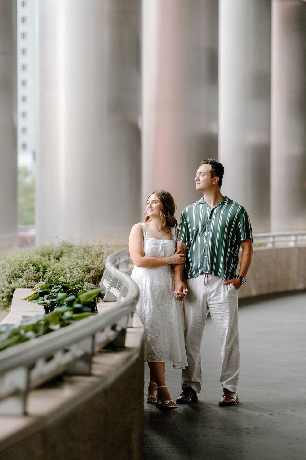 Couple holding hands under large columns in an urban setting, woman in white dress, man in green striped shirt.