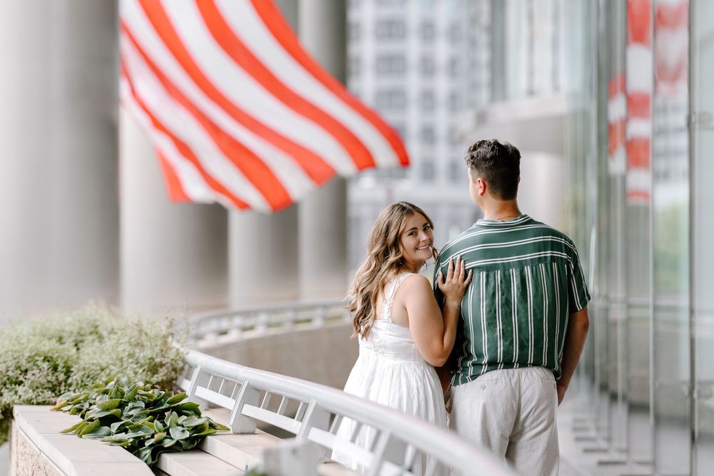 Couple standing on a balcony with an American flag backdrop, woman in white dress smiling at man in green striped shirt.