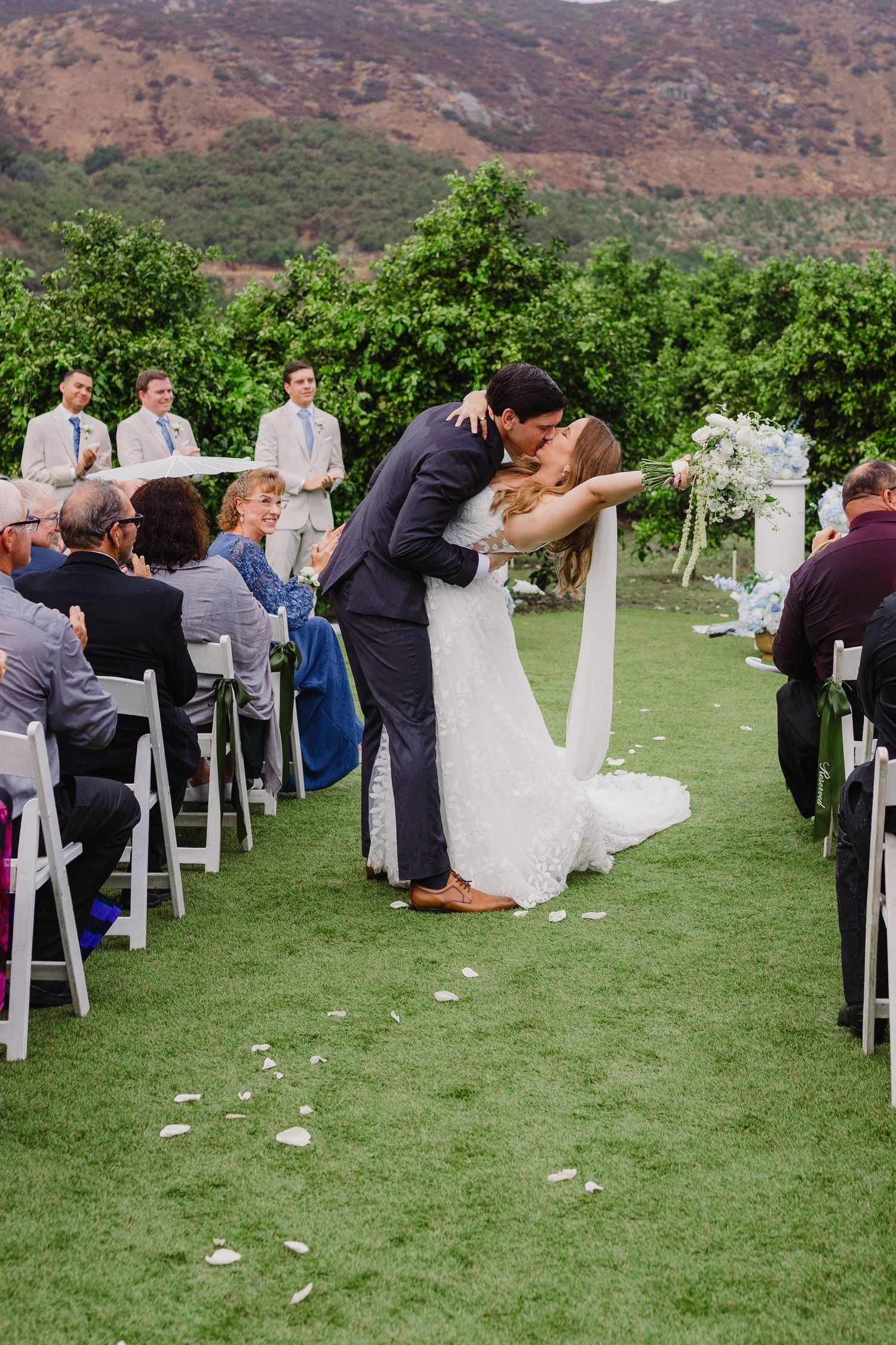 Bride and groom share a dramatic dip kiss at the end of the aisle as seated wedding guests watch at an outdoor ceremony.