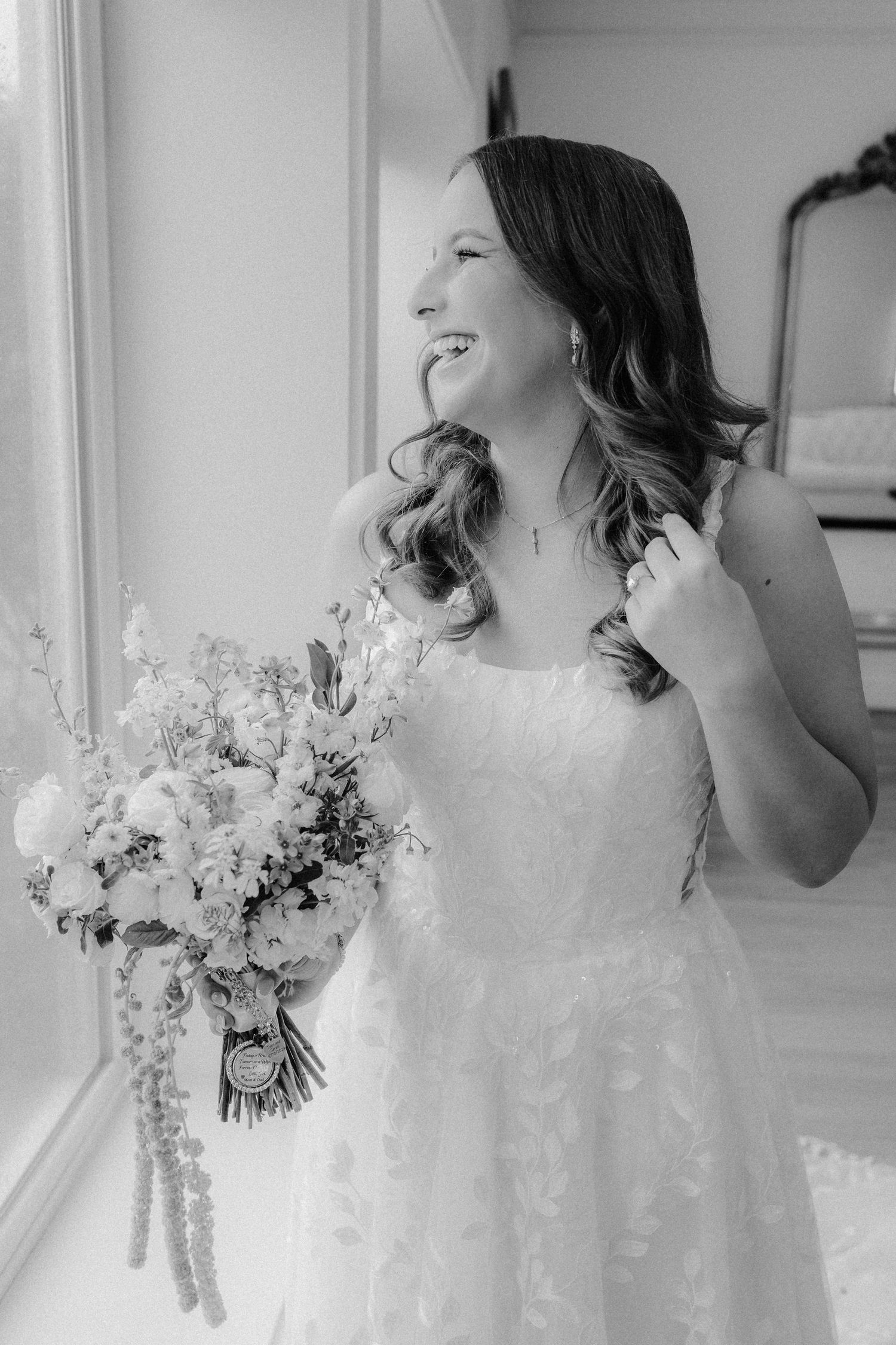 Black and white portrait of a laughing bride in a lace gown holding a bouquet of delicate flowers beside a bright window.