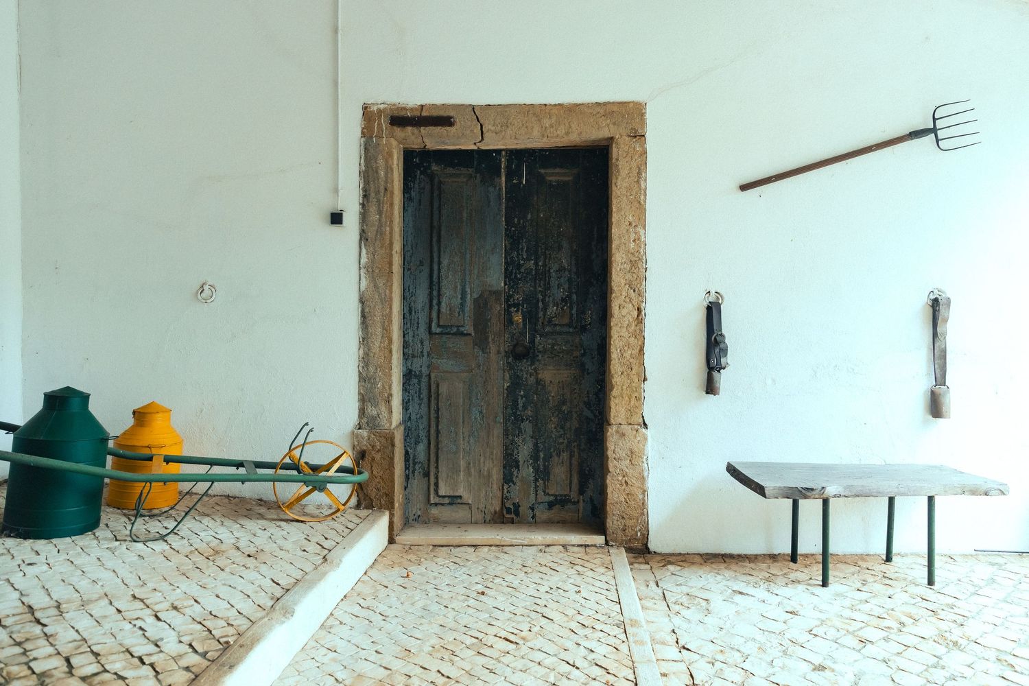Rustic wooden door set in a stone frame on a white wall, with cobblestone floor and vintage farm tools nearby.