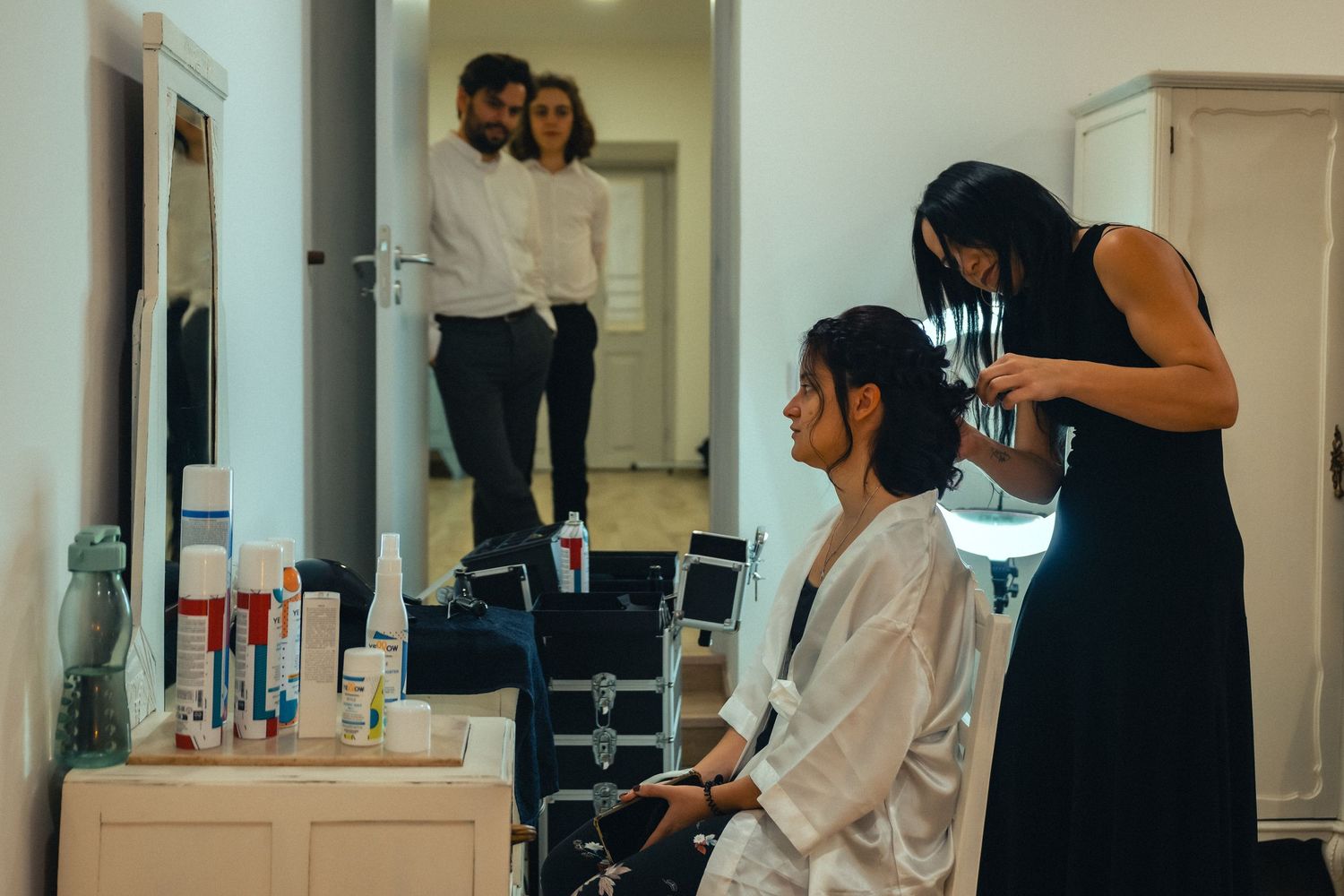 A hairstylist works on a seated bride in a white robe at a vanity mirror while a couple stands reflected in the background.