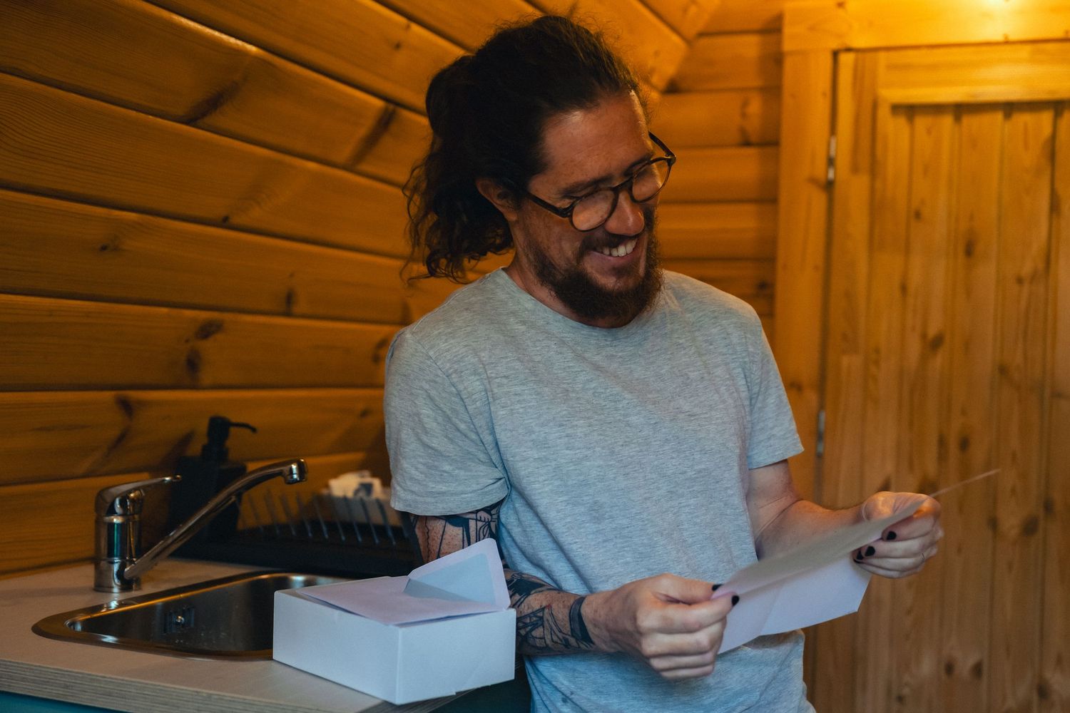 Smiling man with long hair, glasses, and tattoos reading a document in a cozy wooden cabin kitchen.