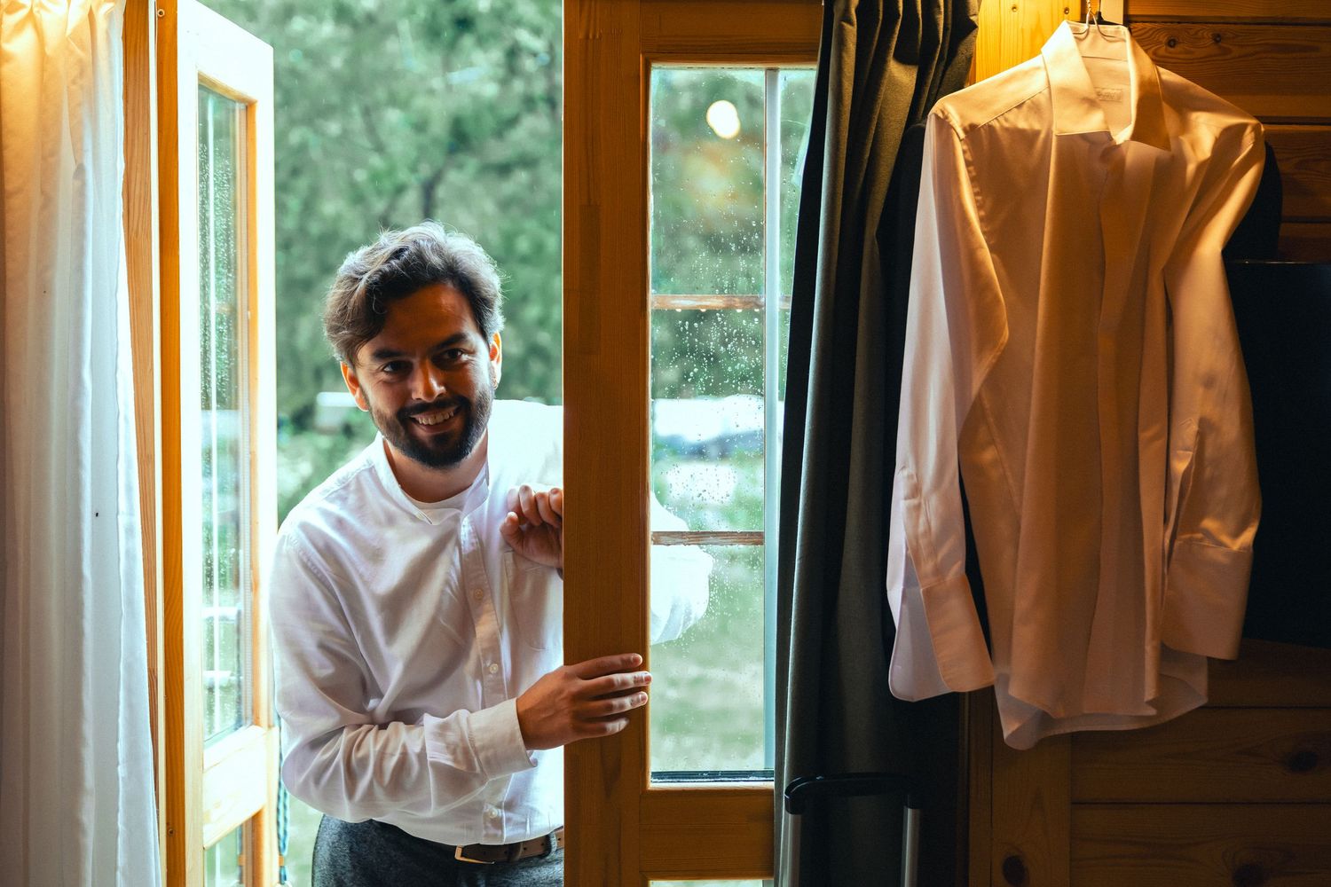 Smiling bearded man in white shirt standing by open wooden door with hanging dress shirts nearby.