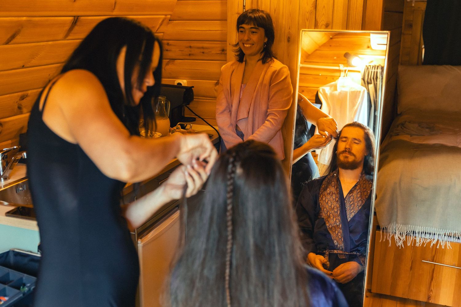 A hairstylist braids a woman's hair in a cozy wooden cabin while two others watch and smile nearby.