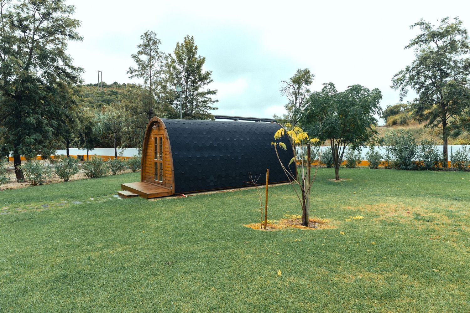 Modern barrel-shaped glamping pod with wooden exterior on lush green lawn near a serene lake surrounded by trees.