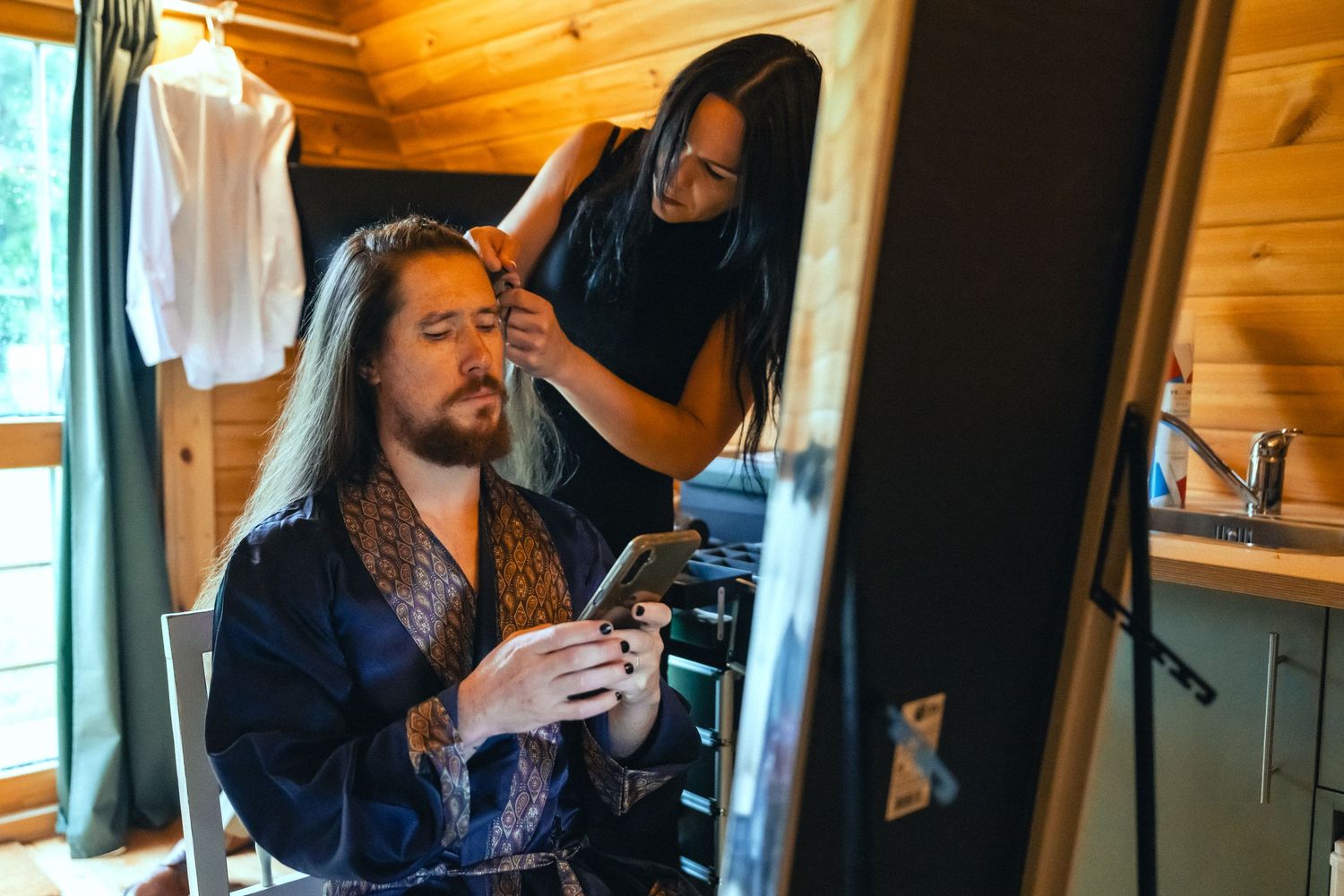Man in robe scrolling phone while woman styles his long hair in a rustic wooden cabin with mirror.