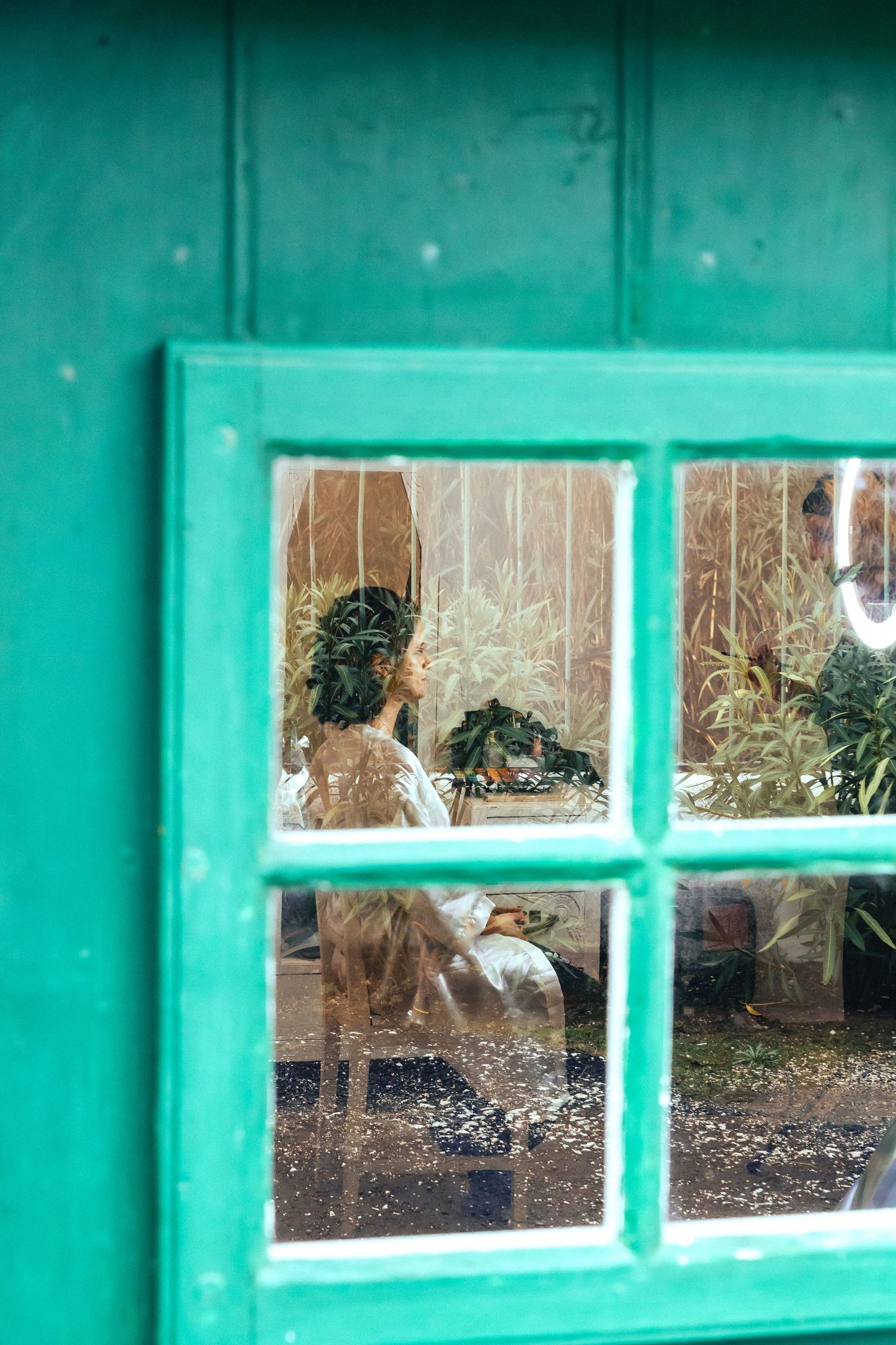 Bride with floral crown sits inside a green-framed window, surrounded by lush plants in a rustic greenhouse setting.