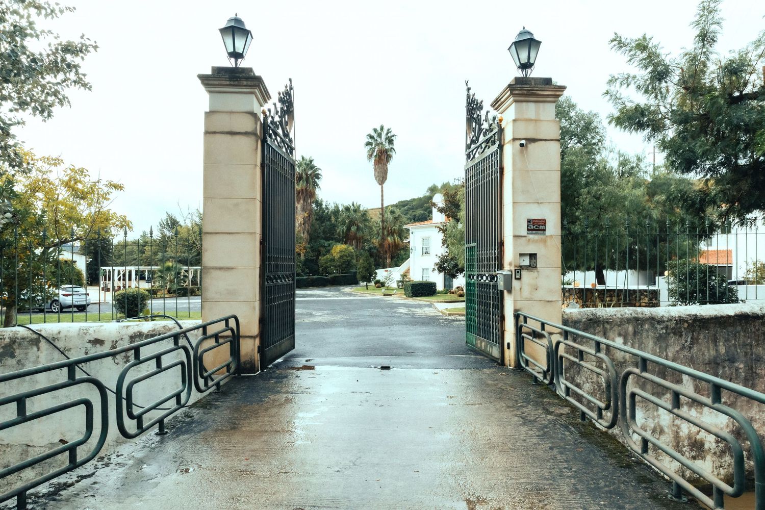 Open wrought iron gates with stone pillars and lamp posts leading to a driveway surrounded by trees.
