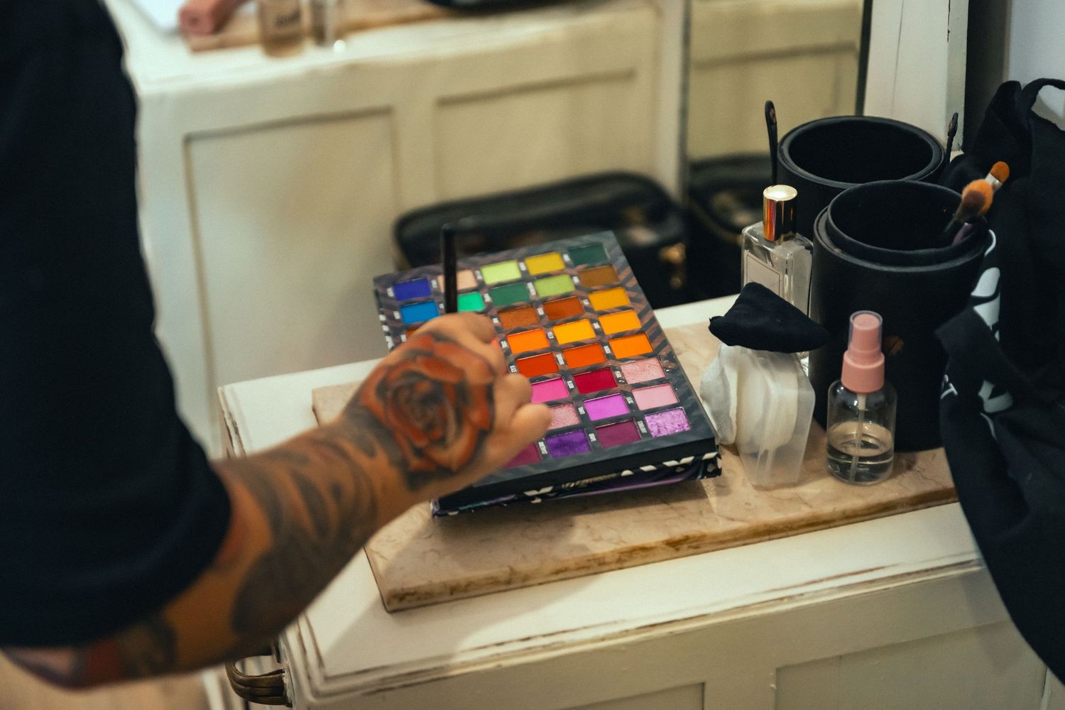 Tattooed hand selecting colors from a vibrant watercolor paint palette on a wooden surface in an art studio.