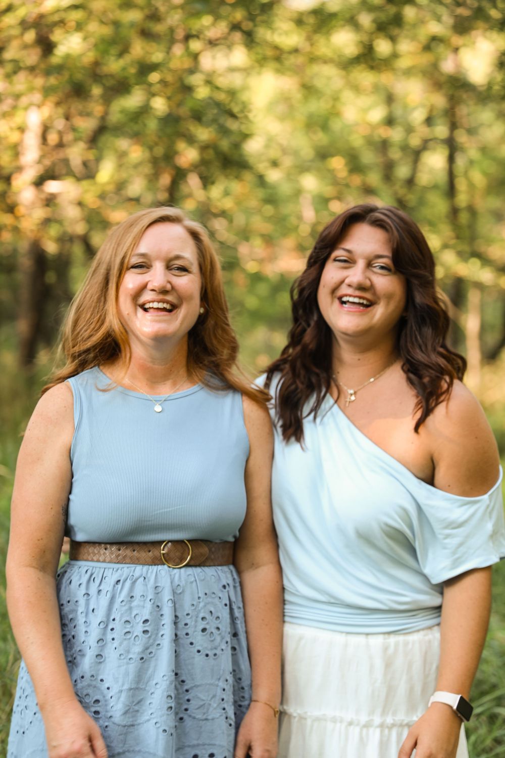 Two smiling women in light blue outfits pose together outdoors in a lush green wooded setting.