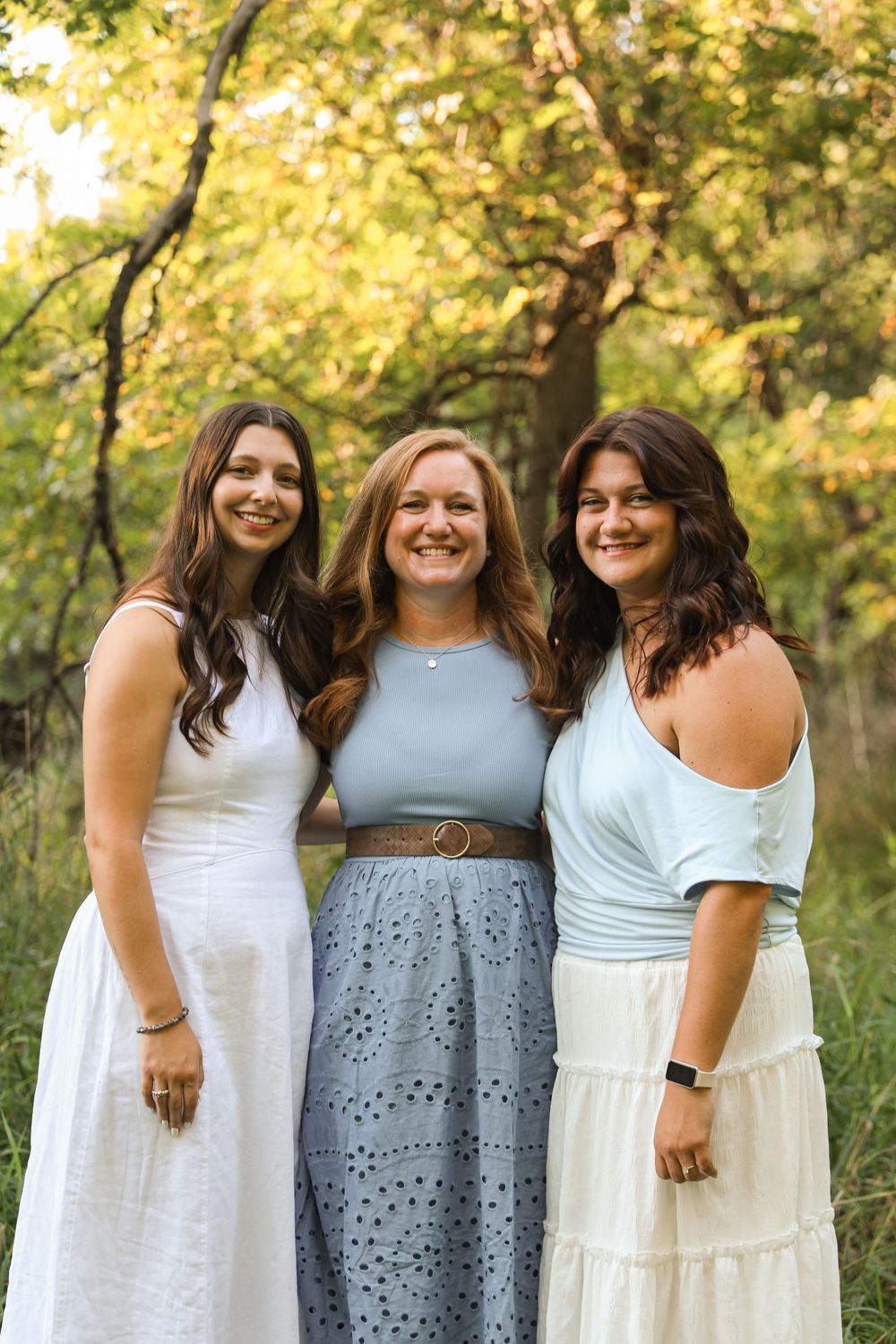 Three smiling young women in light-colored dresses pose together outdoors in a sunlit wooded setting.