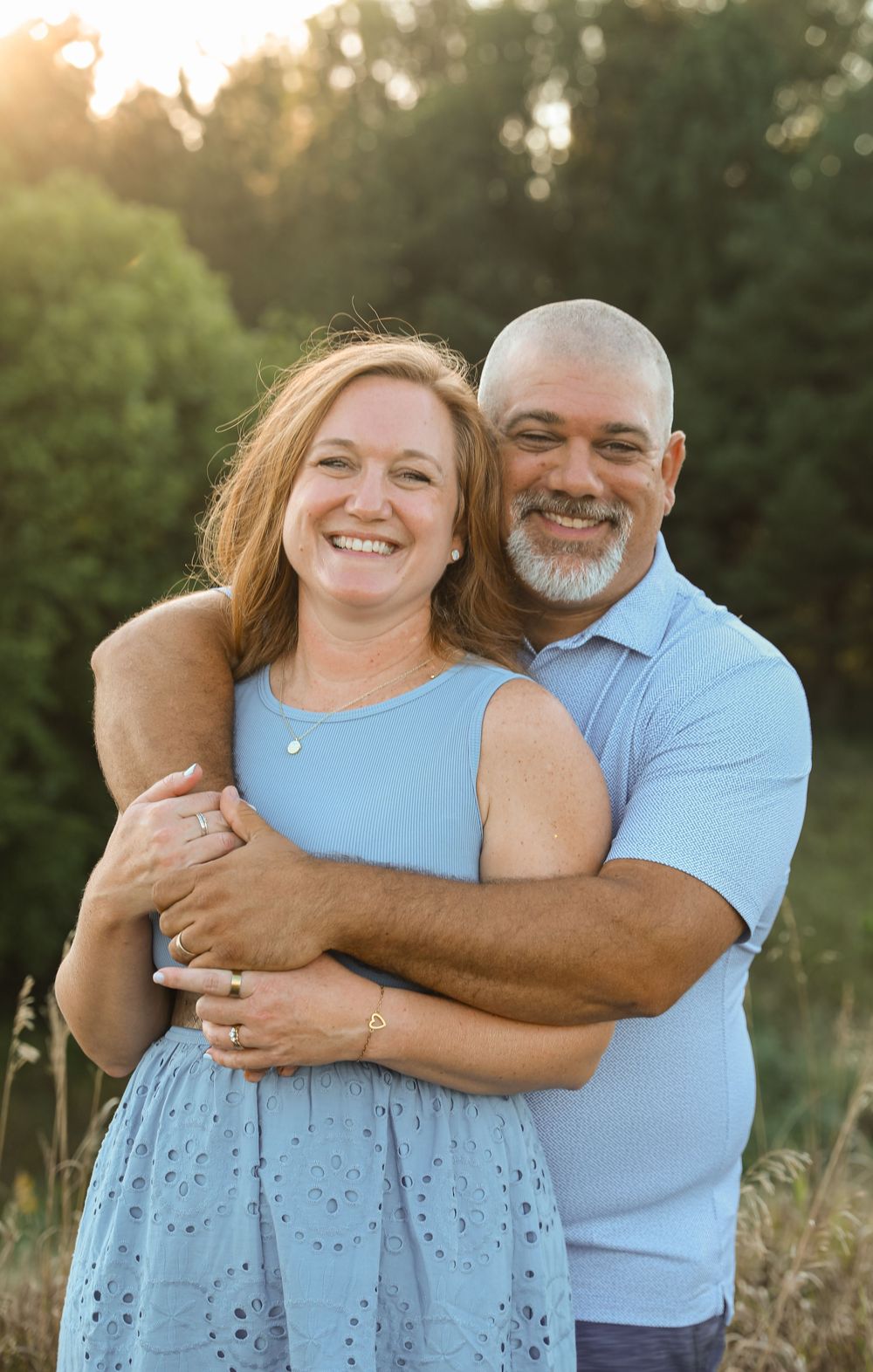 A smiling couple embraces outdoors at golden hour, woman in blue dress, man in light blue polo, with lush greenery behind them.
