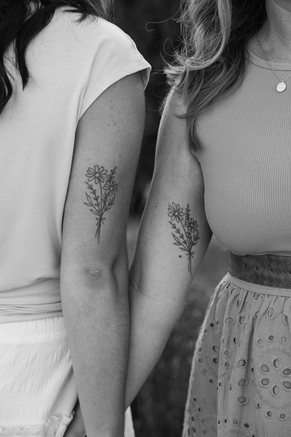 Two women show matching floral bouquet tattoos on their forearms in this black and white photo.