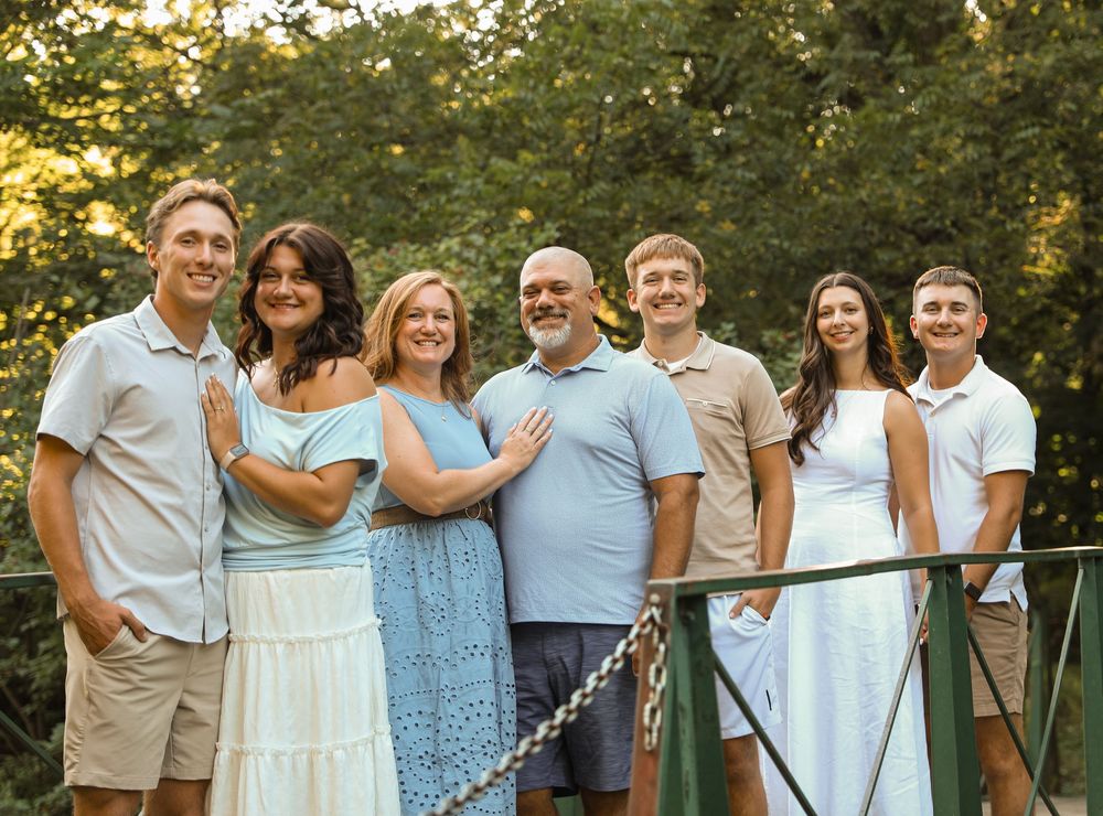 A happy family of seven poses together on a bridge surrounded by lush green trees in warm golden light.