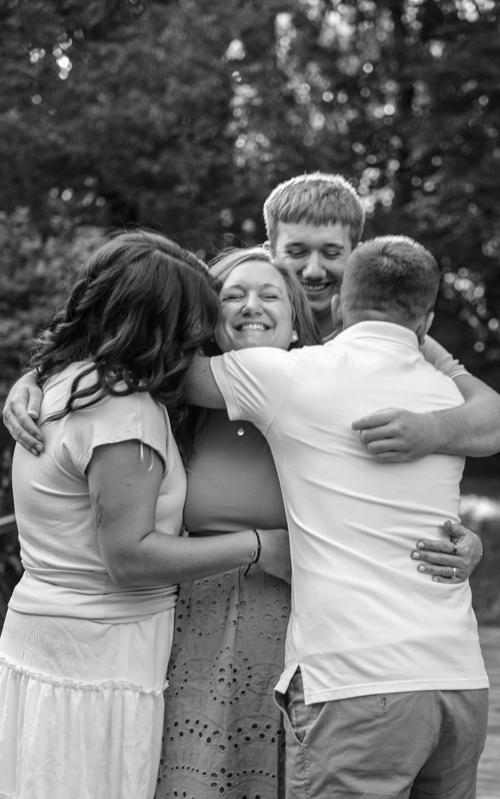 Black and white photo of a joyful family group hug outdoors, smiling woman embraced by three loved ones.