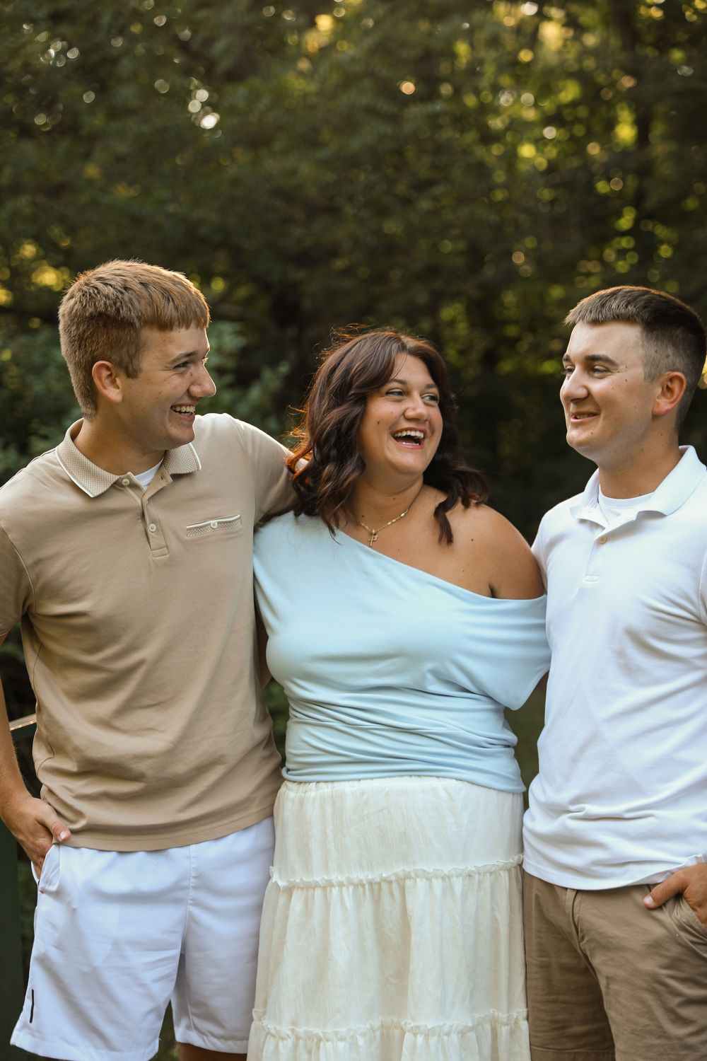 Three young adults smiling and laughing together outdoors, posing in casual summer outfits with green trees behind.