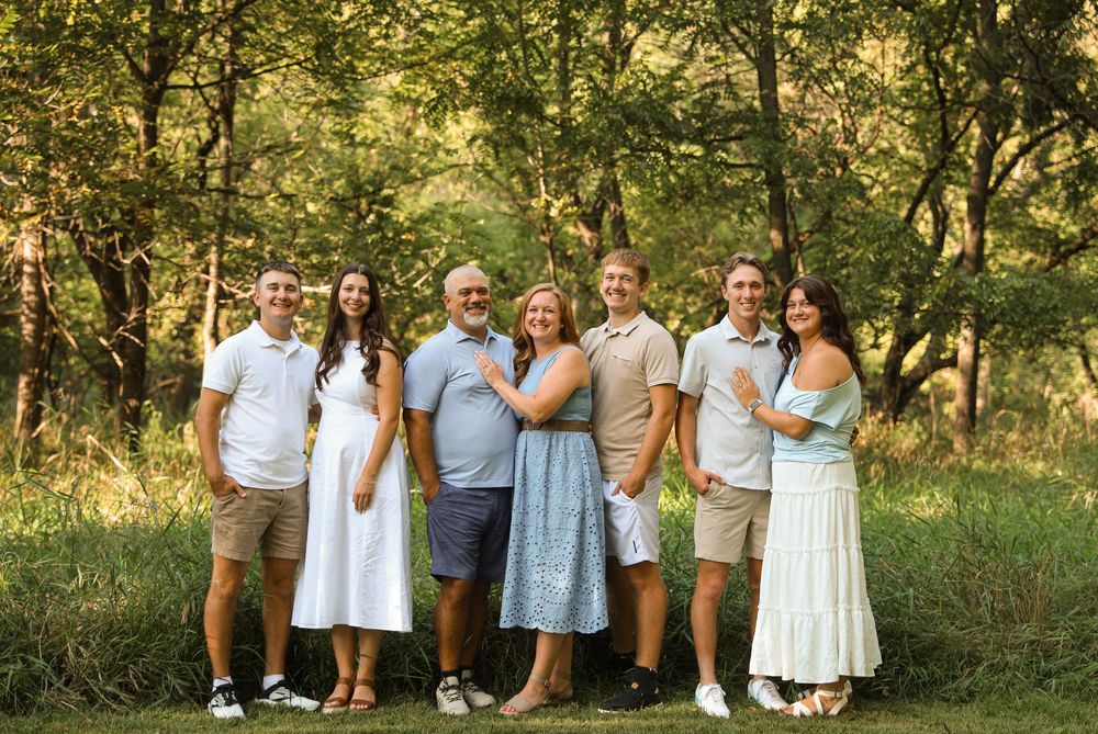 Family of seven smiling outdoors in a lush green park setting, wearing coordinated neutral and pastel outfits.