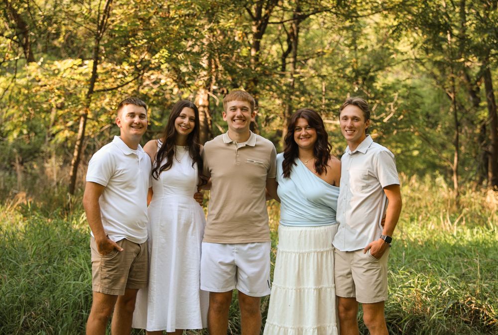 Family of five smiling together outdoors in a lush green forest setting, wearing coordinated neutral and pastel outfits.