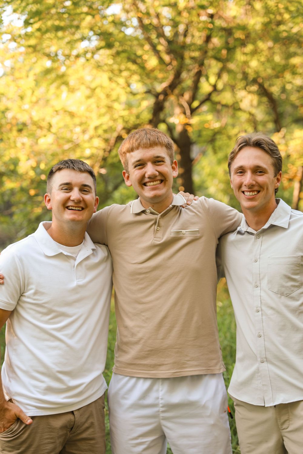 Three smiling young men with arms around each other posing outdoors with green trees in the background.