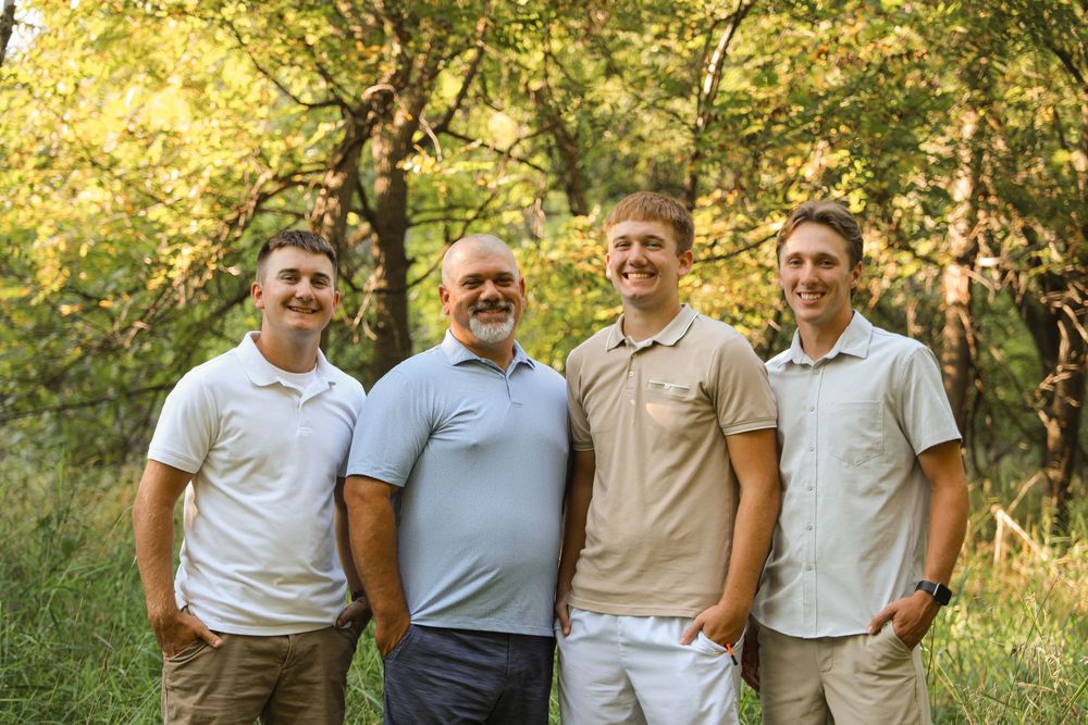 Four smiling men pose together outdoors in a lush green forest setting during a family portrait session.
