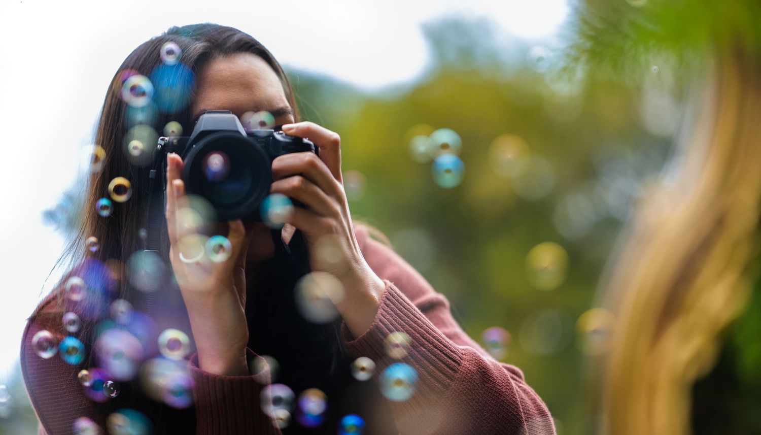 Young woman photographer taking photos with DSLR camera surrounded by colorful soap bubbles.