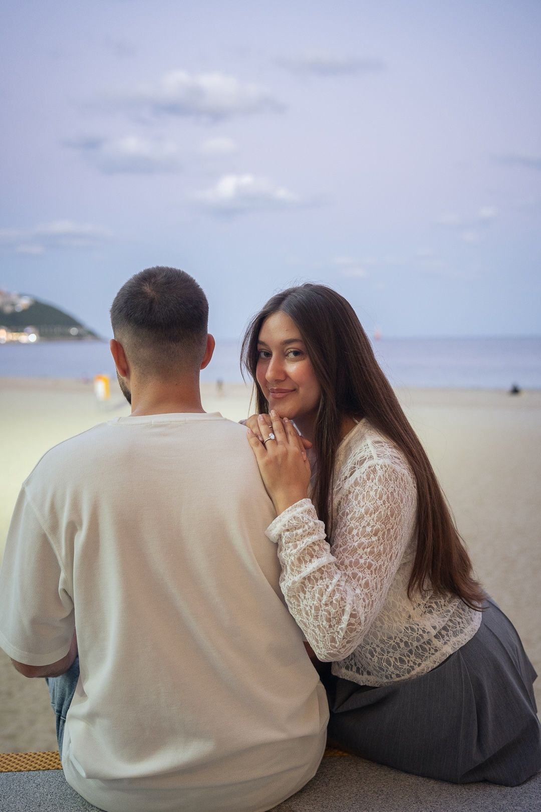 A couple sits on a beach at dusk, woman in white lace top smiles over shoulder showing a ring.