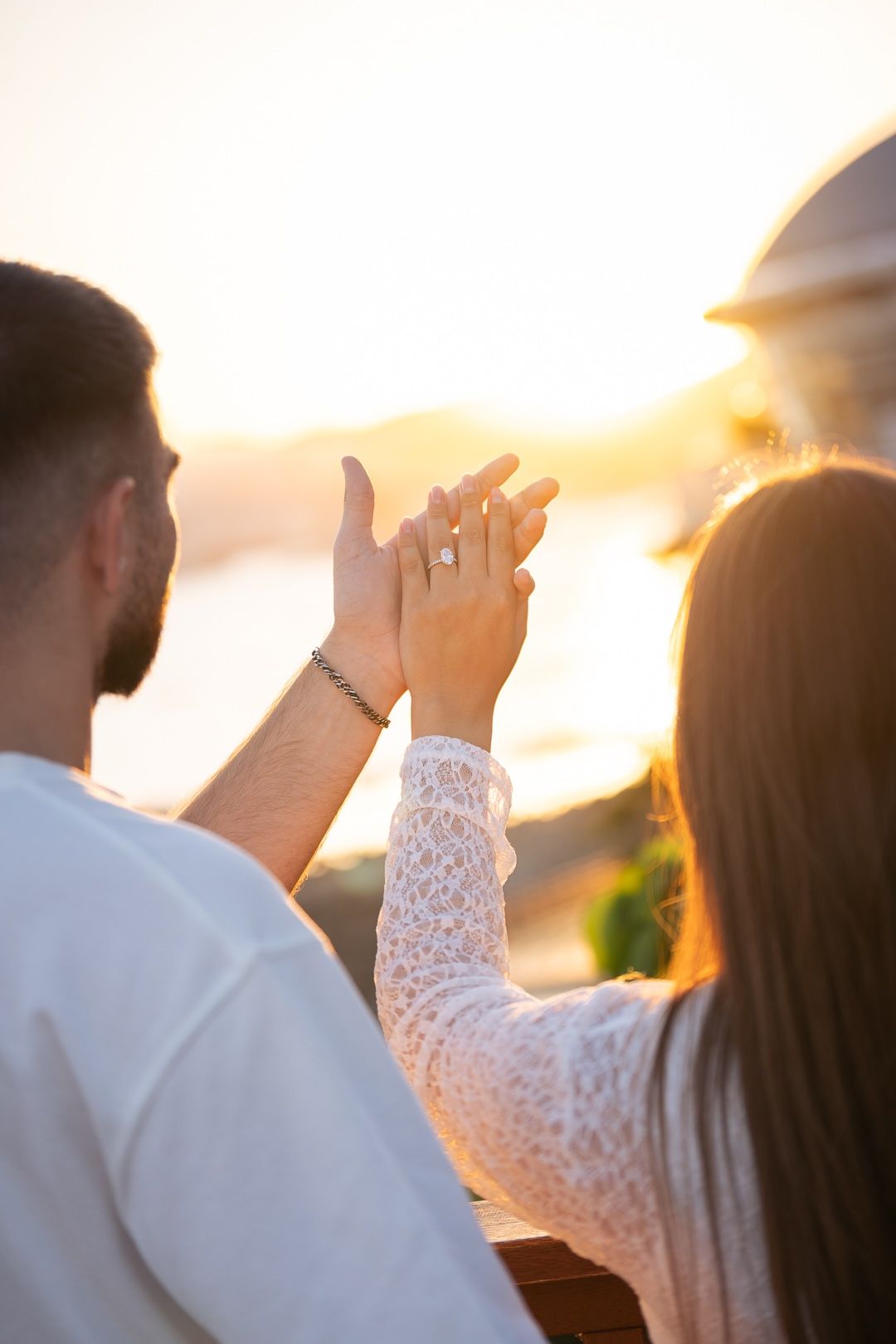 Couple holding hands at sunset showing engagement ring, romantic proposal moment with scenic ocean view backdrop.