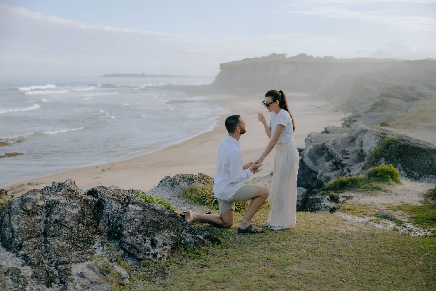 A man kneels on one knee on a rocky coastal cliff, proposing to his partner above a sandy beach and rolling ocean waves.