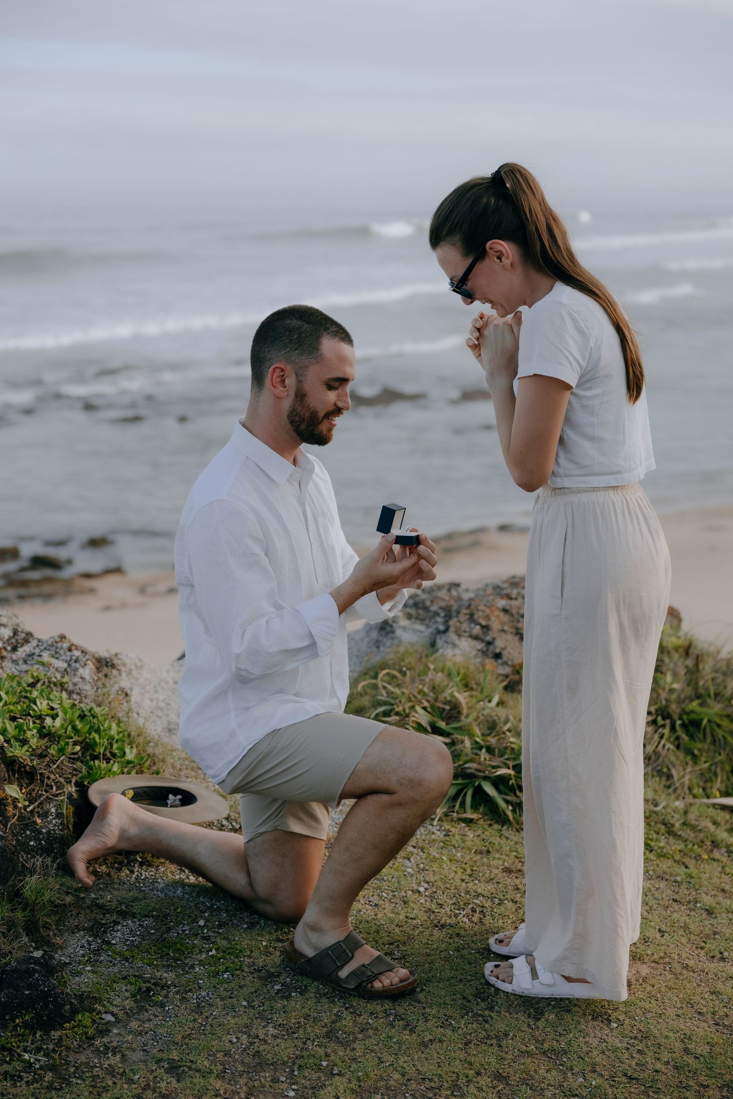 A kneeling man holds out an engagement ring box to his emotional fiancée, who covers her mouth in joy beside the sea.
