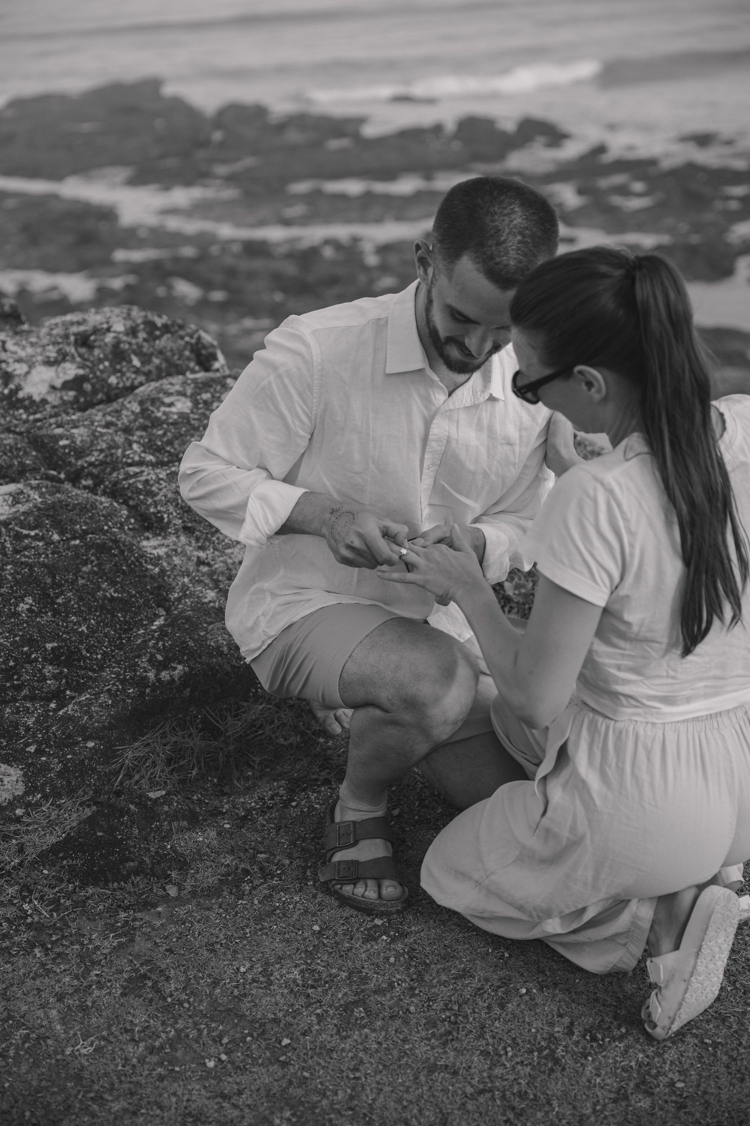 Black and white image of a couple kneeling together on a rocky shoreline as he slips an engagement ring onto her finger.