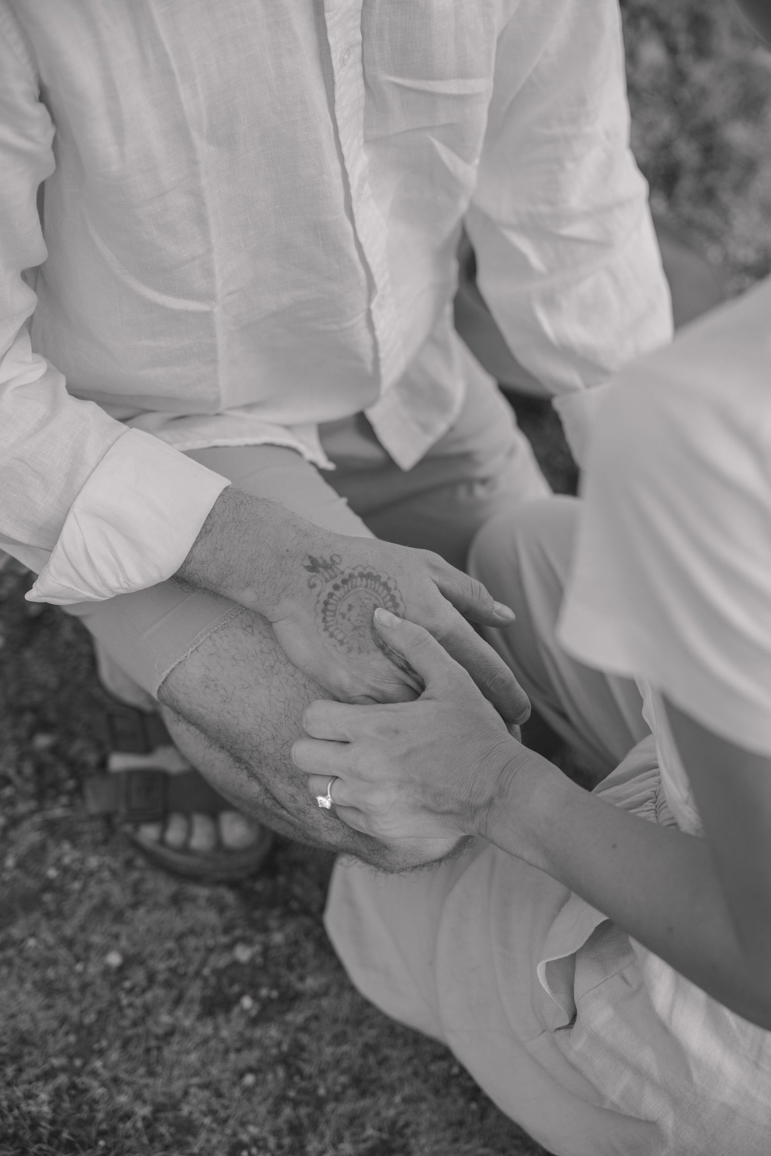Close-up black and white photo of a couple's clasped hands, with an engagement ring on her finger and a tattoo on his hand.