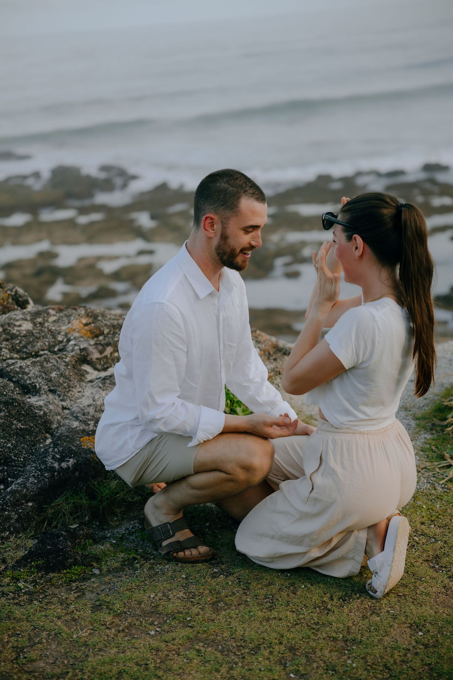 A smiling man kneels before his partner, who wears sunglasses and holds her hand to her face, during a clifftop engagement.