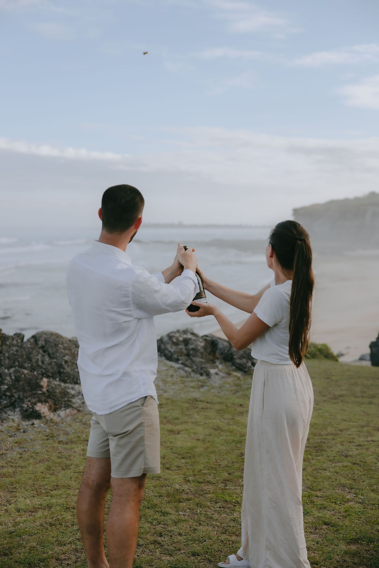 A couple stands on a grassy coastal cliff, celebrating their engagement by opening a champagne bottle above the ocean.