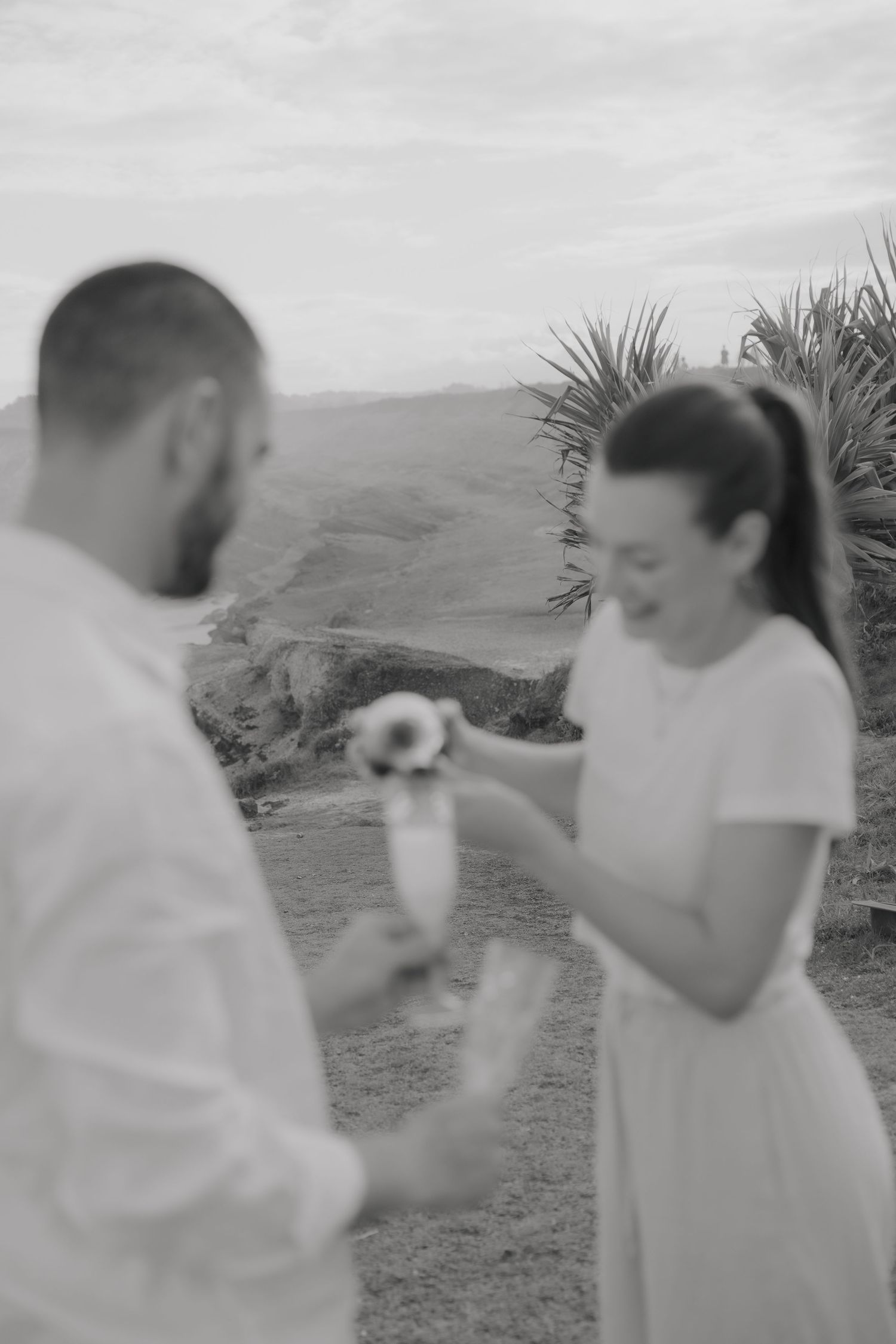 Black and white image of a happy couple laughing while pouring champagne from a bottle into glasses on a scenic hilltop.