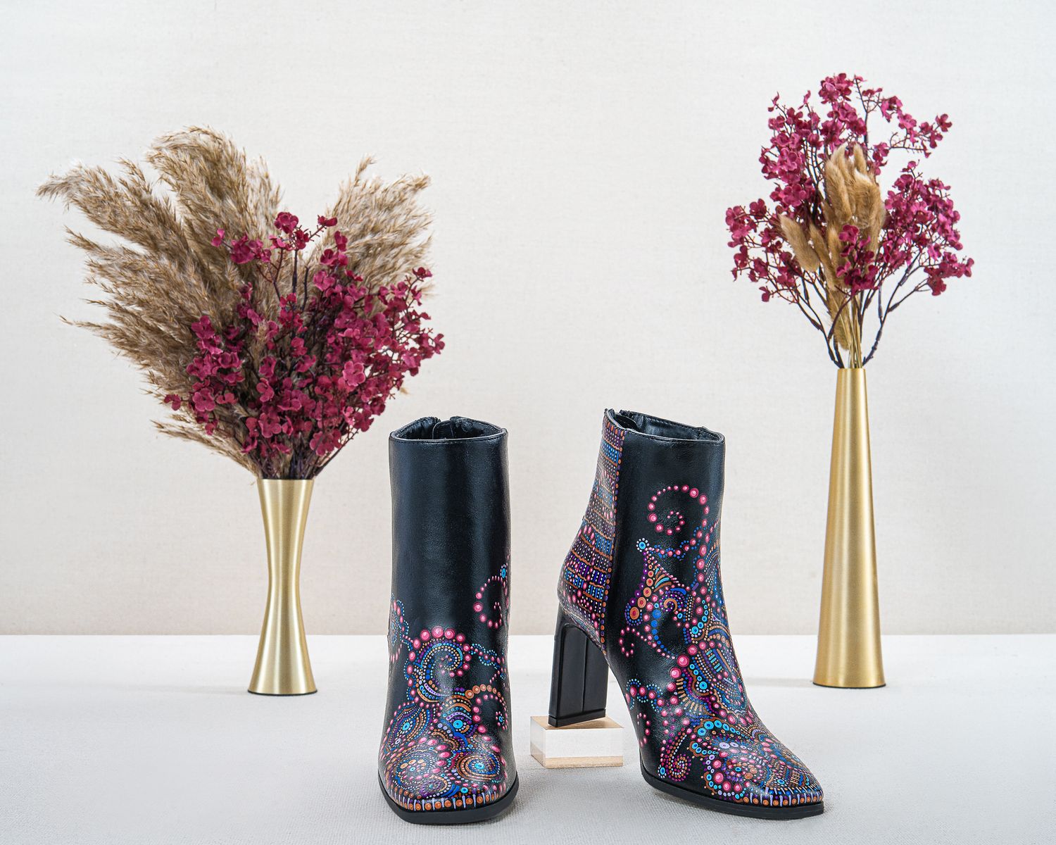 Black leather ankle boots with colorful hand-painted dot art and block heels beside gold vases holding dried pampas grass and pink flowers.
