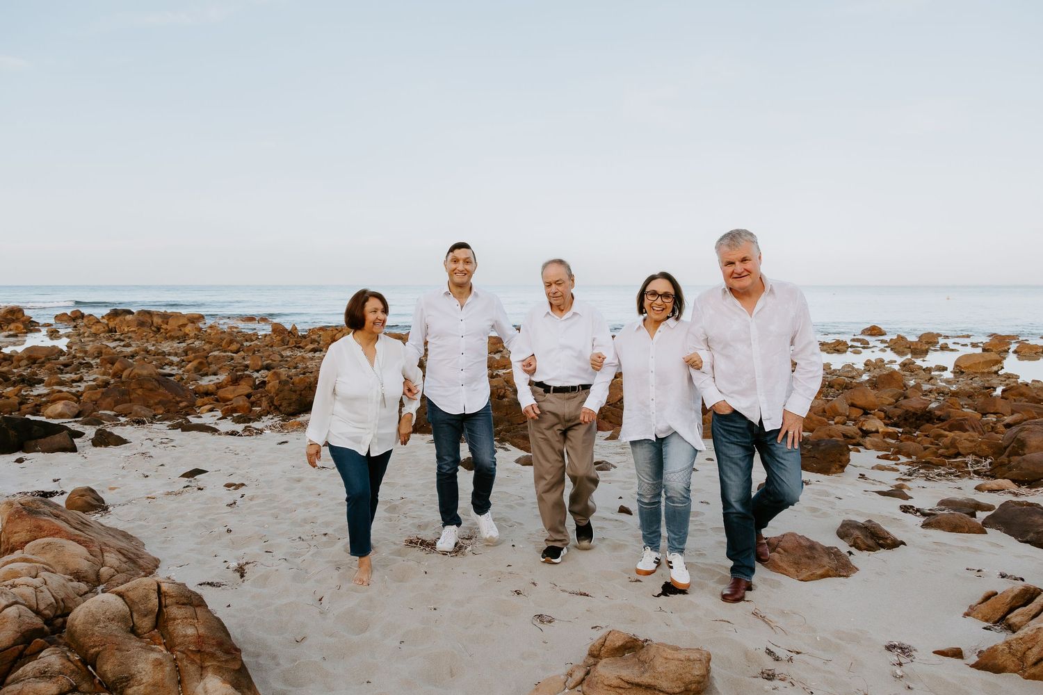 Five people in white shirts and jeans walk arm in arm along a sandy beach dotted with rocks and tide pools under a soft, hazy sky.