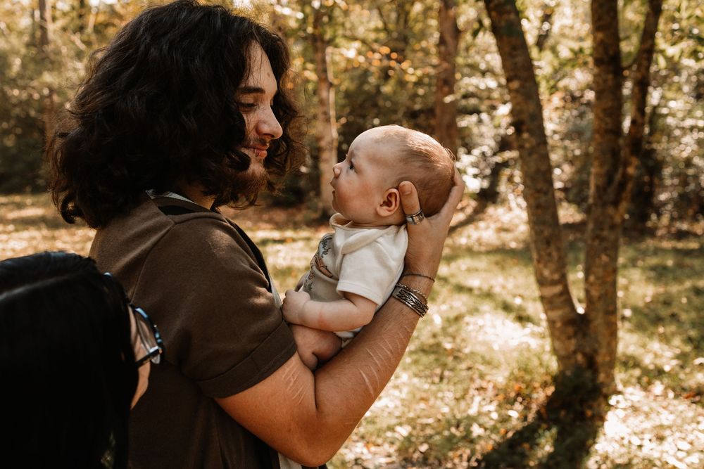 Smiling parent with curly dark hair holds a baby up in the air outdoors among sunlit autumn trees in a peaceful forest setting.