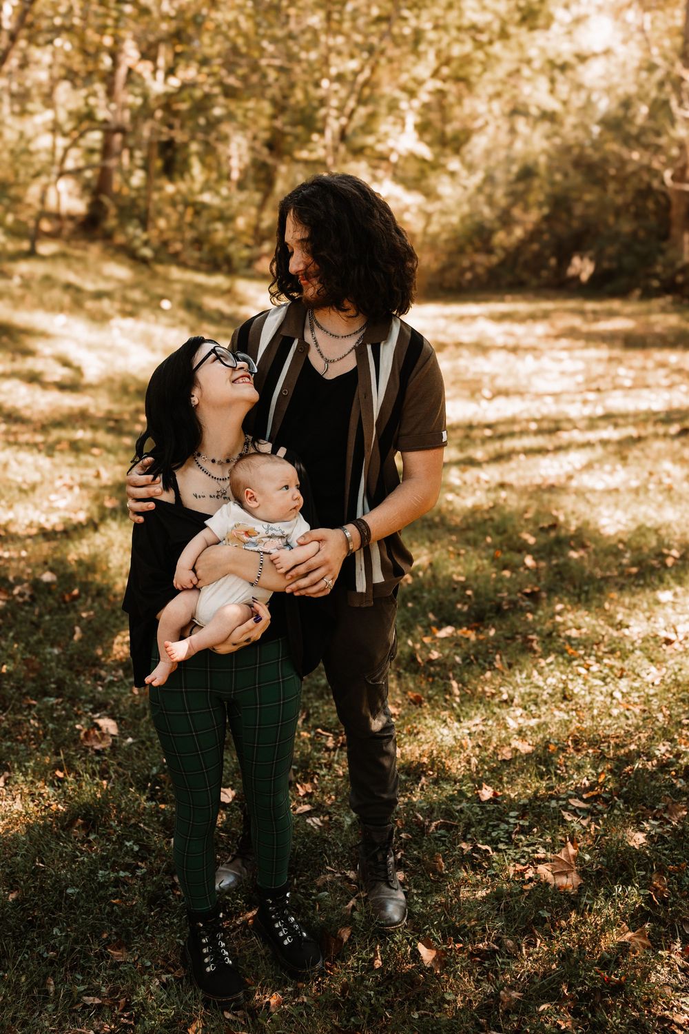 A smiling couple holds their baby while standing on a sunlit grassy path surrounded by autumn trees in a cozy outdoor family portrait
