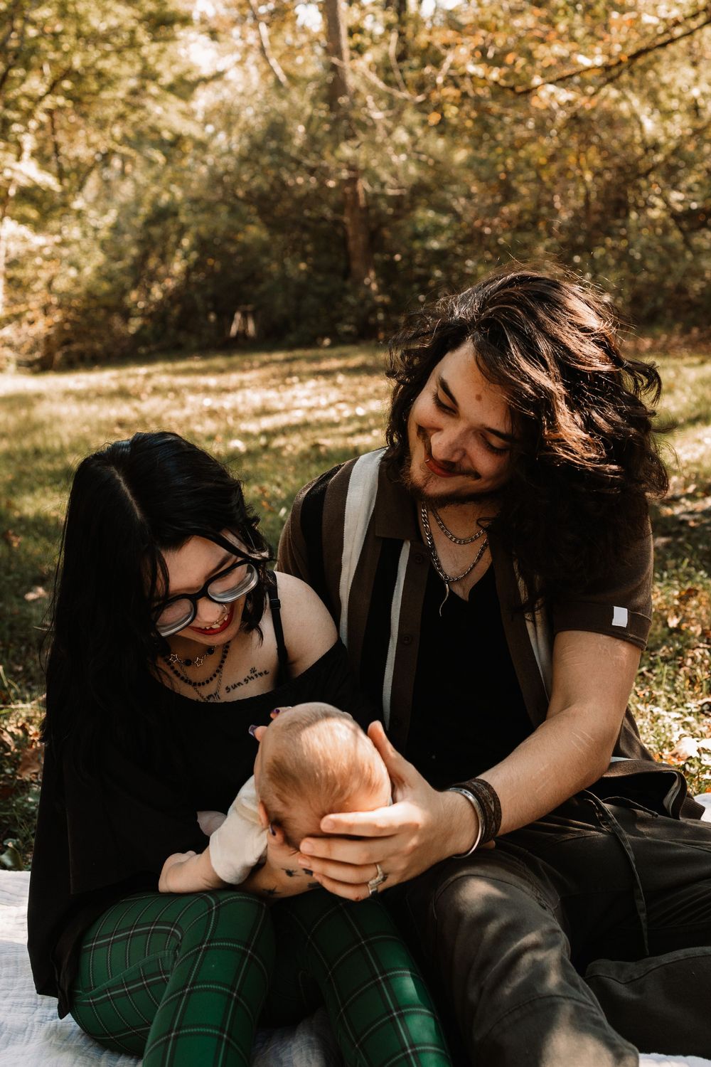 A smiling couple sits on a blanket in a sunlit park with baby during a lifestyle family photography session in Oak Ridge, Tennessee.