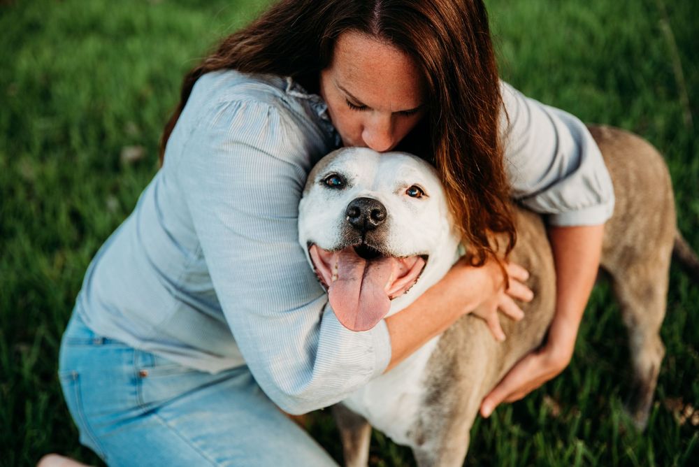 Woman hugging her happy white Labrador dog outdoors on green grass, dog smiling with tongue out.