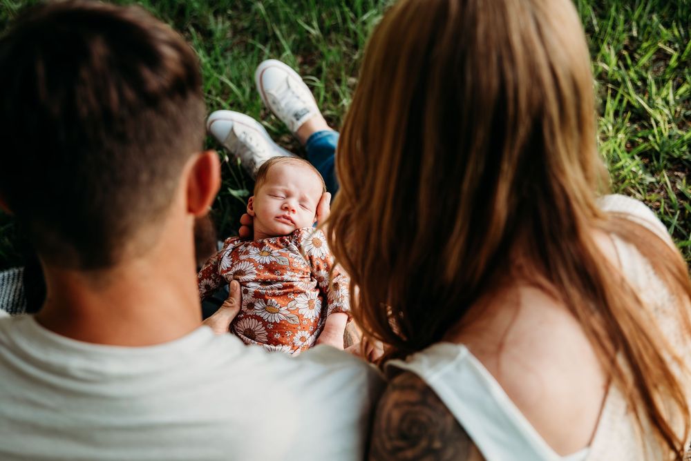 Parents sitting on grass looking down at their sleeping newborn baby dressed in a floral onesie outdoors.