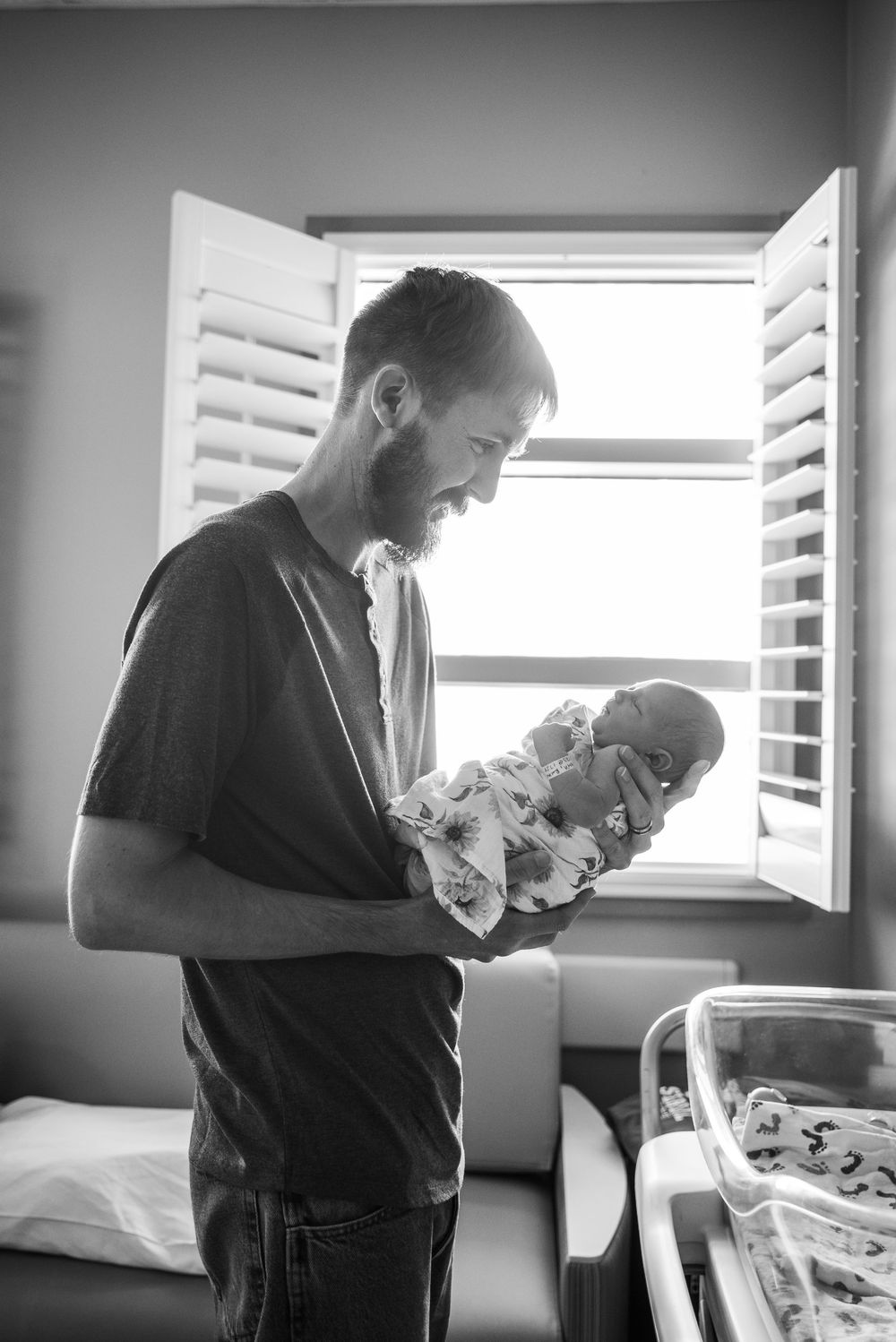 Black and white photo of a bearded father smiling at his newborn baby in a hospital room by a window.
